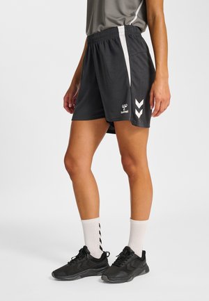 Kobieta ubrana w czarne sportowe spodenki z białym paskiem po bokach i logo w formie jaskółki, do tego białe skarpety i czarne buty sportowe.