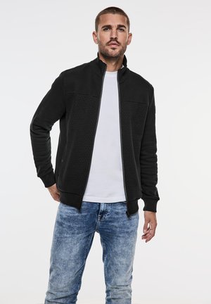 Black zip-up jacket with textured chest panel, ribbed cuffs, and collar. Worn over a white shirt, paired with light blue jeans.
