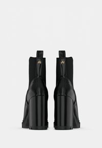 Un par de botines negros de tacón alto con tacones de bloque, paneles laterales elásticos y pequeños detalles en oro en los lazos del tacón, mostrados desde atrás.