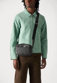 Sort vævet læder crossbody taske med lynlås, iført en model i en mintgrøn suedjakke og brune bukser.