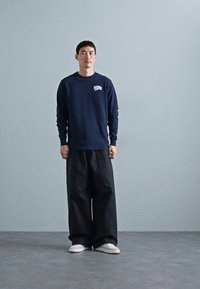 Marineblå sweatshirt med en liten hvit logo på brystet, kombinert med løse svarte bukser og hvite sko, i en minimalistisk innendørs setting.