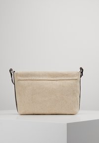 Beige texturerad tygväska med mörkbruna läderremmar, rektangulär form och lockstängning, placerad på en vit yta mot en grå bakgrund.