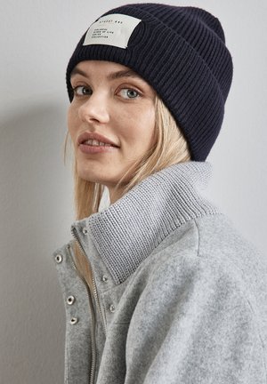 Bonnet en tricot côtelé marine avec une étiquette blanche ; veste zippée grise avec un col haut et texturé ; tissu lisse, accents en matériel métallique.
