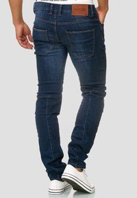 Jeans de mezclilla azul oscuro con un diseño slim-fit, que cuentan con bolsillos traseros y una etiqueta de cuero marrón en la cintura. Acentuados con costuras blancas.