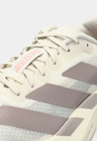 Primo piano di una sneaker Adidas Adizero color crema con strisce e lacci marrone chiaro su sfondo bianco.