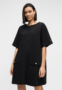 Μαύρο φόρεμα με κοντά μανίκια σε άνετη γραμμή, με δύο εξωτερικές τσέπες και χρυσά κουμπιά. Λείο ύφασμα και στρογγυλή λαιμόκοψη.