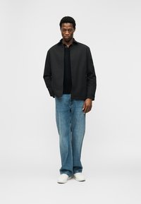 Άνδρας με μαύρο μπουφάν, μαύρο πουκάμισο, μπλε τζιν και λευκά αθλητικά παπούτσια, στάσιμος με το ένα χέρι στην τσέπη, μπροστά σε απλό φόντο.