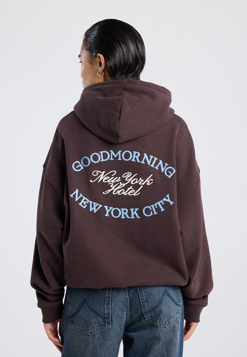 Sudadera con capucha marrón con texto bordado en azul y crema que dice "GOODMORNING New York Hotel" y "NEW YORK CITY" en la parte posterior.