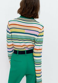 Gestreepte trui in meerdere kleuren met een kraag, gecombineerd met effen groene broeken met een donkere riem. De stof lijkt gestructureerd.