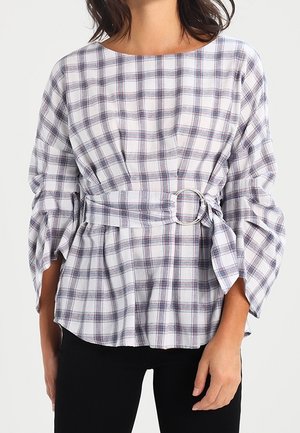 Mujer que lleva una blusa de manga larga a cuadros blancos y azules con un cinturón ancho y hebilla redonda plateada en la cintura, combinada con pantalones negros.