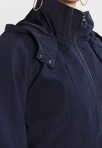 Granatowa, wodoodporna kurtka z wysokim kołnierzem, zapięciem na zamek błyskawiczny, akcentami na ramionach oraz detalami zapinanymi na napy. Gładka tekstura, bez widocznych wzorów.