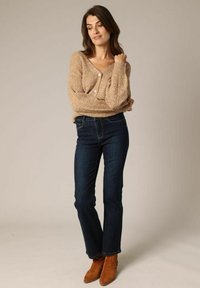 Beige gebreide cardigan met een V-hals en knoopsluiting, gecombineerd met donkere blauwe spijkerbroek en tan enkellaarsjes.