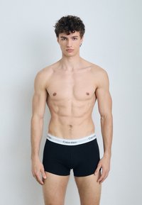 Boxers noirs en coton avec une ceinture élastique blanche portant le logo "Calvin Klein". Le modèle est torse nu et se tient debout.