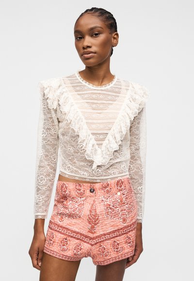 Jeune femme aux cheveux tressés portant un chemisier blanc en dentelle avec des volants et un short taille haute rose à motifs, debout devant un fond uni.