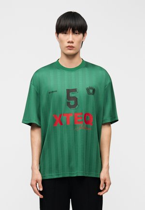 Młody mężczyzna o prostych czarnych włosach, ubrany w luźną zieloną sportową koszulkę w paski z numerem 5 i czerwonym napisem "XTEQ".