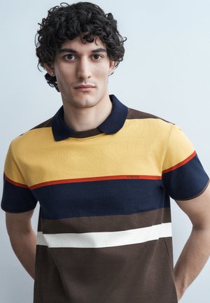 Fiatal férfi göndör, sötét hajjal, mustárszínű, tengerészkék, fehér és barna csíkos galléros pólót visel, és közvetlenül a kamerába néz.