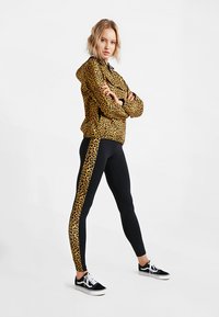Leopardprint, ritsjack gecombineerd met zwarte leggings met een leopardstreep. Model draagt zwarte sneakers en heeft de armen gekruist.