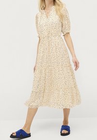 Blommig midiklänning i ljus tan, med puffärmar, elastisk midja och lagerdesign. Kombinerad med blåa slidesandaler och en mjuk textur.