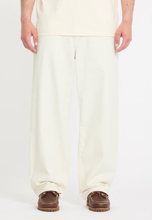 Personne portant un pantalon ample de couleur blanc cassé et des mocassins en cuir marron, debout devant un fond blanc uni.