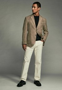 Blazer en daim marron clair avec des revers notés, fermeture à un bouton, poches latérales, associé à une chemise noire et un pantalon écru, chaussures noires.