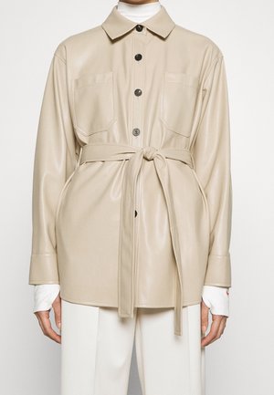 Chaqueta de cuero beige con botones, dos bolsillos en el pecho y un cinturón atado, llevada sobre un jersey de cuello alto blanco y pantalones blancos.