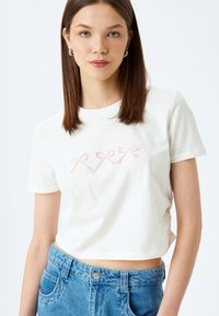 Witte cropped t-shirt van katoen, met drie roze geborduurde strikken op de voorkant. Gecombineerd met hoge blauwe denim jeans.