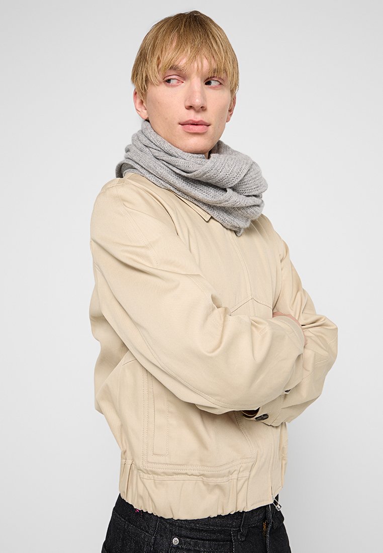 Chaqueta bomber beige con puños elásticos, combinada con una bufanda de punto gris claro envuelta alrededor del cuello, exhibiendo un diseño casual.