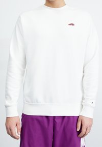 Hombre vestido con un sudadera blanca lisa con un pequeño parche de zapatilla roja en el pecho y pantalones cortos morados de Nike, frente a un fondo blanco.