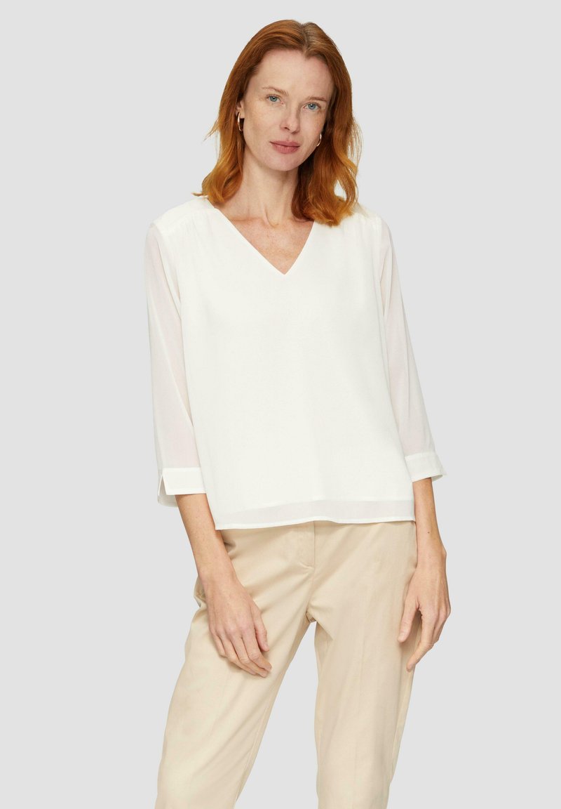 Blouse blanche à col en V avec des manches trois-quarts, accents en tissu transparent aux manches, associée à un pantalon beige clair. Texture lisse, design minimaliste.