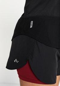 Czarne sportowe szorty z warstwową konstrukcją, wyposażone w siateczkową podszewkę i czerwoną warstwę pod spodem. Charakterystyczne akcenty z logo i brandingiem w kolorze szarym.