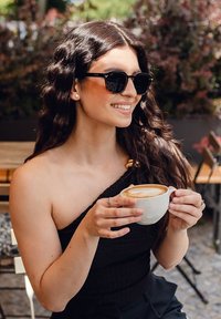 Černé sluneční brýle s oranžovým tónem, top na jedno rameno v černé barvě, držící bílý hrníček s latte artem, dlouhéOndřejské vlnité vlasy, venkovní kavárenské prostředí.