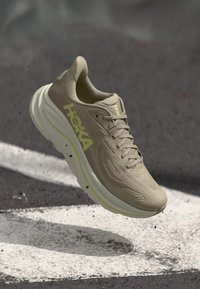 Beige athletic shoe with a padded collar, mesh upper, yellow accents, and a thick, cushioned white midsole, designed for comfort and support.