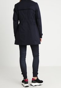 Námořnická modrá parka s úzkým pasem, knoflíkovými manžetami a zadním rozparkem. Nositá přes tmavé džíny a sportovní boty s růžovými akcenty.