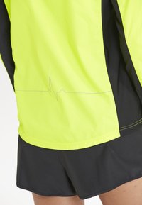 Gelbe, leichte Sportjacke mit einem Herzschlagmuster, kombiniert mit schwarzen Shorts. Der Stoff hat eine glatte Textur und kontrastierende Nähte.