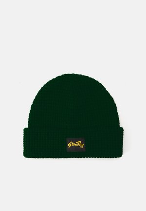 Berretto lavorato a maglia verde con una texture a coste, dotato di una toppa nera con testo giallo. Il cappello ha un risvolto per una vestibilità aderente.