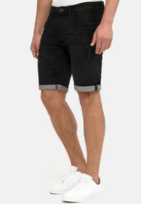Schwarze Jeans-Shorts mit umgeschlagenen Säumen, fünf Taschen, schlankem Schnitt und dezenten Steppnähten. Kombiniert mit weißen Sneakern.