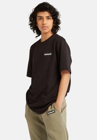 Svart bomull t-skjorte med korte ermer, med en liten hvit Timberland-logo på brystet. Båret med olivengrønne joggebukser.