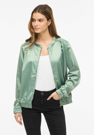 Ung kvinde med lyst brunt hår iført en skinnende mintgrøn bomberjakke, hvid top og sorte jeans, står med den ene hånd i lommen.