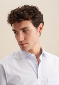 Camicia button-up azzurro chiaro con un sottile motivo a pois, dotata di colletto, taschino sul petto e una morbida texture del tessuto. Modello che guarda leggermente in basso.