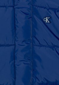 Tmavomodrá prešívaná páperová bunda s prešívanou textúrou, s malým vyšitým logom CK. Hladký nylonový materiál so lesklým povrchom.
