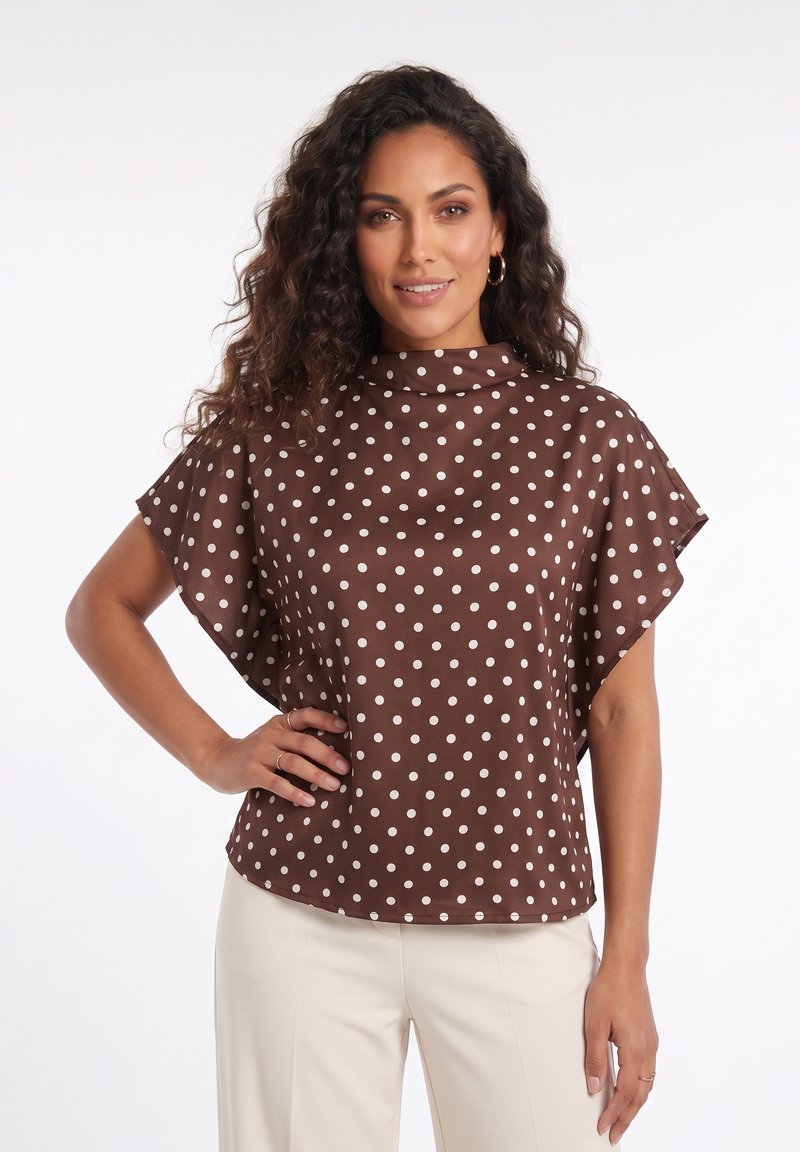 Femme aux longs cheveux bouclés portant un haut marron à pois et un pantalon crème, souriante avec une main sur la hanche sur fond blanc.