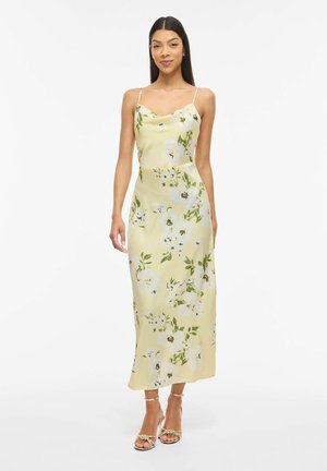 Kvinde iført en gul blomsterprintet slipkjole med spaghetti-stropper og guldfarvede, remmehælede sandaler, stående mod en hvid baggrund.