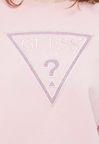 Camisa rosa con un logo triangular de Guess en purpura pastel brillante, que presenta un gran signo de interrogación y las palabras "Washed Jeans" en el frente.