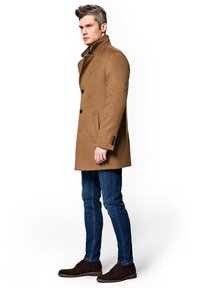 Brązowy płaszcz z mieszaniny wełny, z wysokim kołnierzem, zapięciem na pojedyncze guziki i kieszeniami po bokach, w zestawieniu z niebieskimi dżinsami i ciemnobrązowymi butami.