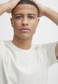 Hvid bomulds T-shirt med rund halsudskæring, afslappet pasform og glat tekstur. Modellens hænder er hvilende på hovedet, med en neutral baggrund.