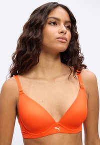 Νεαρή γυναίκα με μακριά κυματιστά μαλλιά που φοράει πορτοκαλί σουτιέν Puma, κοιτάζοντας ελαφρώς προς τα πάνω και προς τα πλάγια, με φόντο απλό.