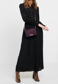 Abito lungo nero con bottoni davanti, maniche lunghe e design plissettato. Abbinato a una borsa a tracolla viola e stivali con tacco a fantasia.