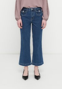 Μπλε τζιν καμπάνα με ψηλή μέση, δύο μπροστινές τσέπες και άβαφη γαζιά στο τελείωμα. Συνδυασμένο με μαύρες γόβες μυτερής μύτης.