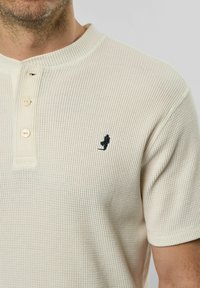 Kremfarget polo-skjorte med en teksturert, ribbet mønster. Har en knappestolpe og en liten mørk logo på brystet. Korte ermer.