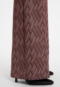 Wijd uitlopende broek van een bruine stof met een geometrisch patroon van zwarte en roze strepen. Zwarte puntige hakschoenen zichtbaar aan de onderkant.